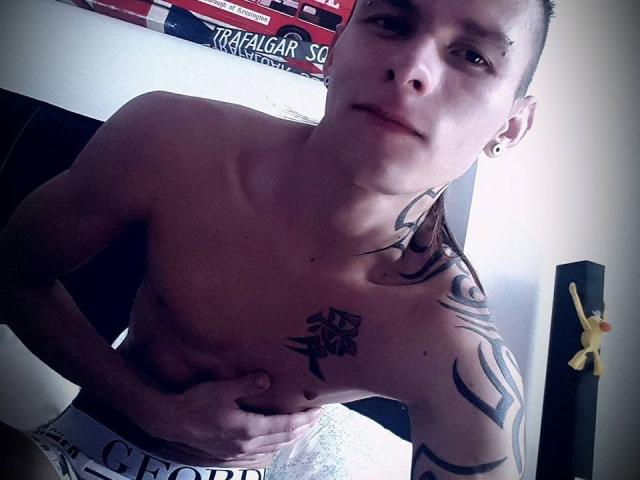 YeremyWalker, sin camisa, con tatuajes, sujetándose las nalgas, cabello castaño corto, aro pequeño en la nariz, perforación en la oreja, mirando a la cámara, con una ligera sonrisa, piel clara, tatuaje tribal negro en el pecho..