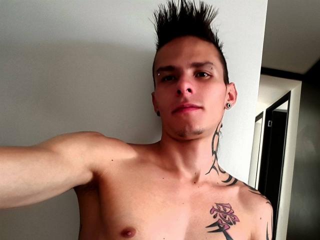 Yeremy Walker, sin camisa, con cabello negro y puntiagudo, pezón pequeño, tatuajes en el pecho y el cuello, perforaciones en las orejas, expresión seria, de pie en una habitación con una puerta en el fondo..
