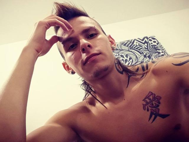 YeremyWalker com um corte de cabelo estilo punk, sem camisa, com tatuagens no peito e ombro, alargadores de orelha pretos, mão direita tocando a cabeça, cabelo castanho levemente bagunçado, expressão séria, iluminação suave..