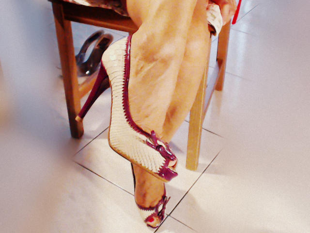 EtoileMilf's pale, wrinkled legs in white high heels with purple accents, crossed at the ankle, resting on a wooden chair with shiny black shoes underneath.