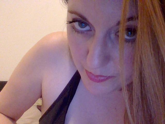 FrenchLea with blue eyes, pink lipstick, black strap, and blonde hair, looking up at the camera with a slight smile, showing her bare shoulders and cleavage.