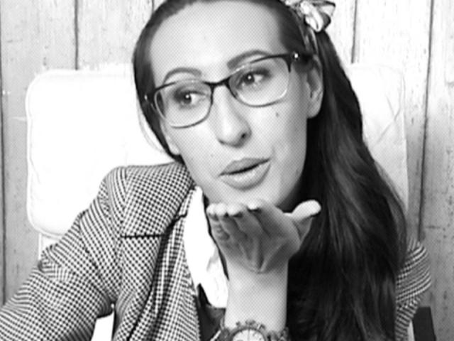 LysAddiction with glasses, checking her nails, wearing a checkered jacket, and a hairbow, looking slightly annoyed. Her tits are not visible, but her face is. Black and white.