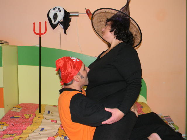 Keyx, con un cappello da strega, un vestito nero e la pelle abbronzata, sta ricevendo un bacio da Keyx, che indossa una maglietta arancione, una bandana rossa e maniche nere, e che le sta toccando il sedere..