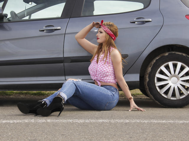 AmyPure sitting on pavement, wearing pink checkered top, blue jeans, black high heels, red bandana, touching forehead, gray car in background, long red hair, medium-sized tits, fair skin.