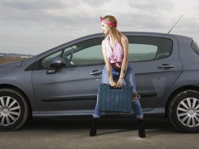 AmyPure with red hair, pink checkered top, blue jeans, black boots, holding a blue suitcase, standing in front of a gray car, cloudy sky, overcast day, looking left.
