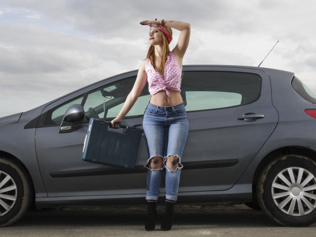 AmyPure leans against a gray car, wearing a pink tie-dye top, blue ripped jeans, black boots, and a red headscarf, holding a blue suitcase, arm raised.