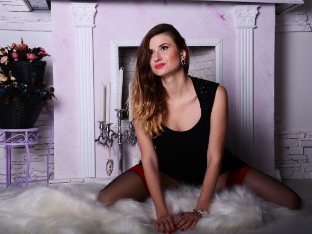 SweetLipsJenny with long blonde hair, black sleeveless top, red underwear, black stockings, and red lipstick, kneeling on white fur, hands on floor, looking at camera, smiling.