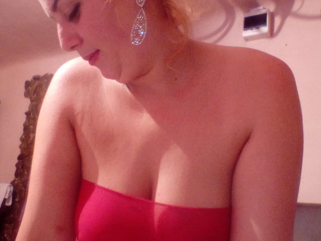 GrosSeinsKatty con un vestido rojo sin tirantes, mostrando sus pechos, con aretes colgantes, mirando hacia abajo con una ligera sonrisa, cabello rubio en un moño despeinado, piel clara, en una luz suave..