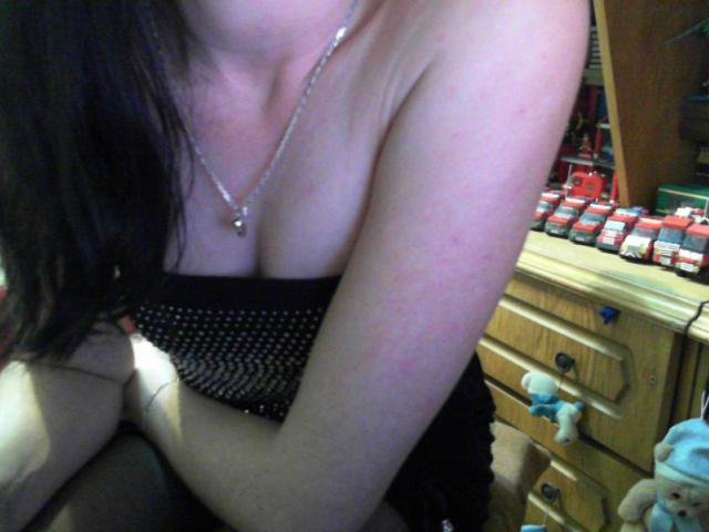 1KinkyBrunette with bare shoulders, black mesh top, small silver necklace, and visible boobs, sitting with arms crossed, in a dimly lit room. Her black hair is partially visible.
