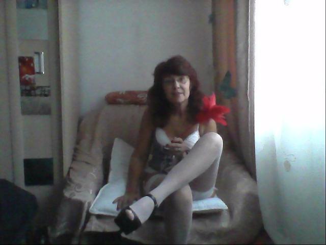 Ceryane, met rood haar, zit op een beige stoel, gekleed in witte lingerie, een korset en witte kniekousen, terwijl ze een rode veer vasthoudt en haar achterwerk laat zien..
