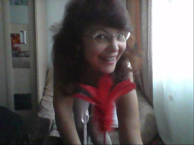 Ceryane with curly brown hair, glasses, and a red feather in her chest, smiling at the camera, wearing nothing but the feather. Her tits are partially visible.