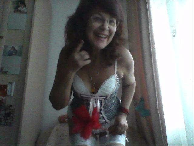 Ceryane, avec ses cheveux bruns bouclés, souriante, pointant du doigt, portant un soutien-gorge blanc avec des rubans rouges et roses, et un pantalon blanc déchiré, se tenant dans une pièce faiblement éclairée..
