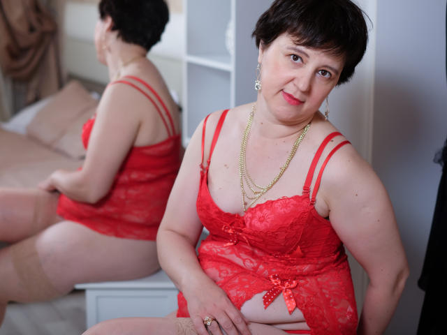 ChristaRose, a chubby woman with short black hair, wearing red lace lingerie, a gold necklace, and beige stockings, sits smiling in a mirror's reflection. Her tits and ass are visible.