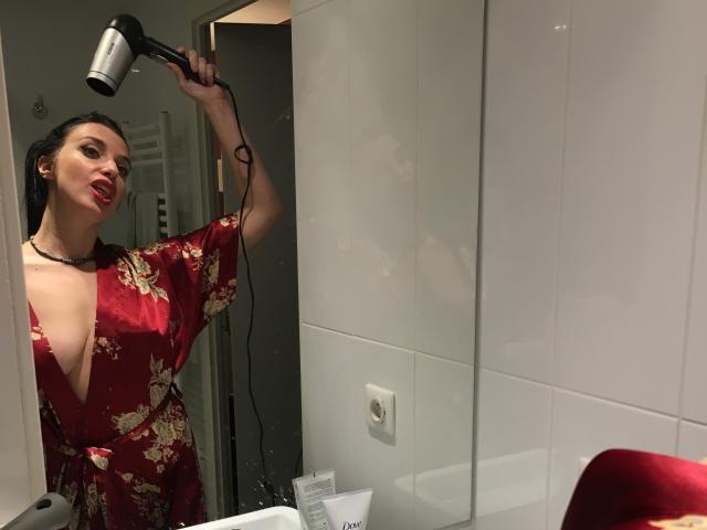 KatieFrenchie with dark hair, red floral robe, deep cleavage, holding a hairdryer, smiling, mirror reflection, white tiled bathroom, red chair bottom right.