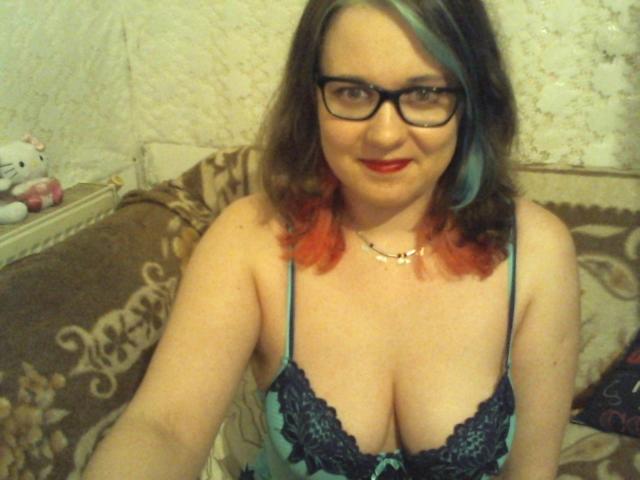 ChaudeBella69 met een bril, rode lippenstift en blauw-groen haar met oranje punten, draagt een kantachtig turquoise en zwarte beha, toont haar grote borsten en glimlacht naar de camera..