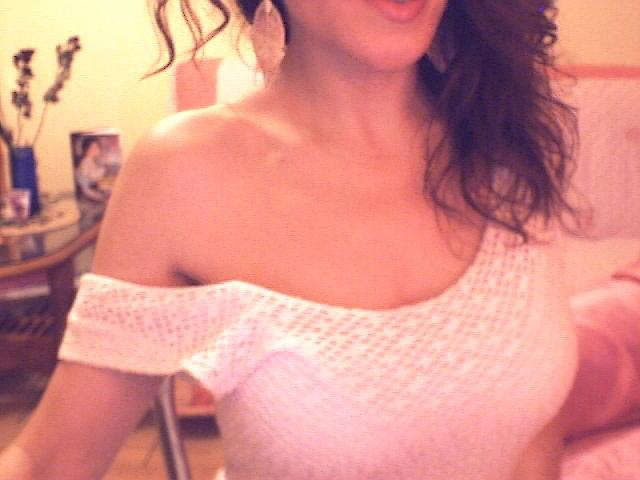 Venusya with wavy brown hair, wearing a white off-shoulder knit top, showing her small tits and collarbone, holding a pink pillow. Soft, warm lighting.