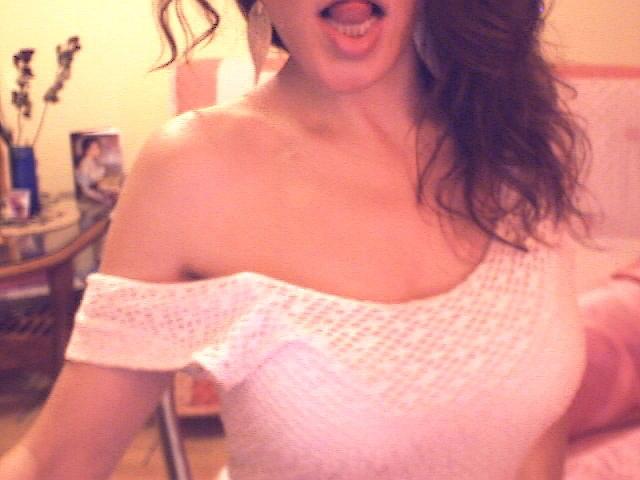 Venusya with wavy brown hair, tongue out, wearing a white off-shoulder knit top, showing her boobs. Soft, warm light, slightly blurred, creating a cute, casual vibe.