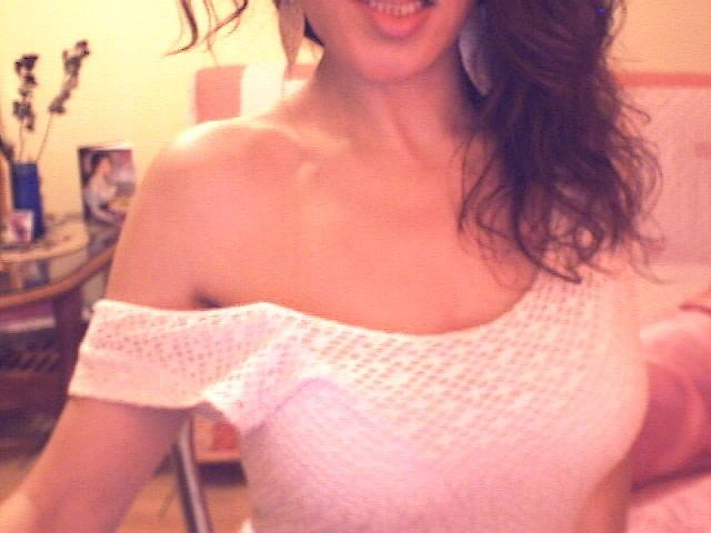 Venusya with wavy brown hair, smiling, wearing a white off-shoulder knit top, showing her tits. Soft pink bedroom in background. Warm, slightly blurred lighting.