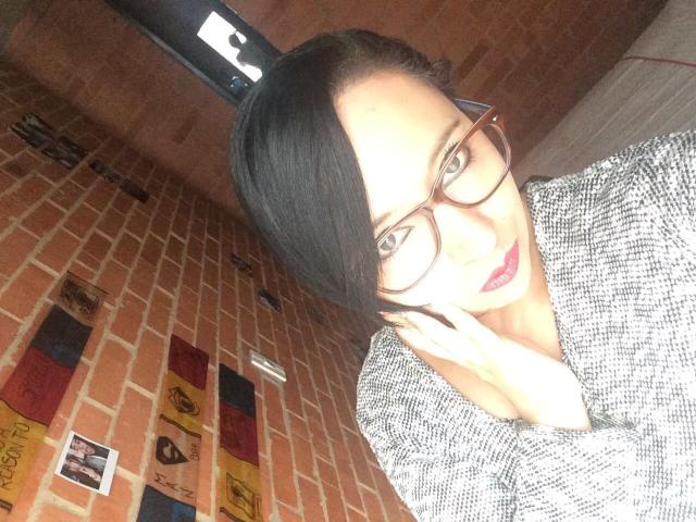 BellaLolita with glasses, black hair in a side ponytail, gray textured shirt, holding phone to ear, red lipstick, brick wall, overhead light, slightly tilted head.