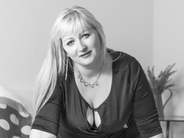MarinaSweet, blonde with bangs, smiling, wearing a dark, cleavage-revealing top with a necklace, has medium tits, and is leaning forward in a black-and-white photo.