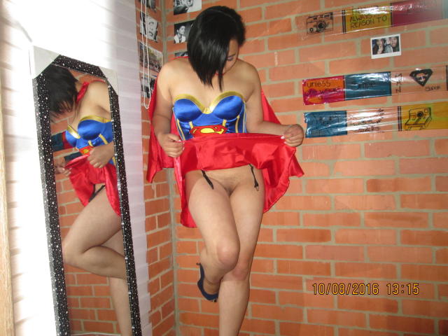 BellaLolita with black hair, blue and yellow Supergirl costume, red skirt, and bare pussy, lifting skirt, standing in front of mirror, brick wall, and posters, dated 10/09/2013.