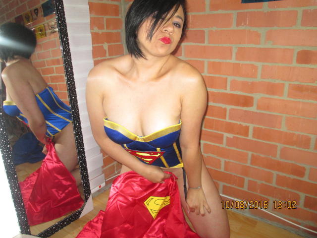 BellaLolita con i capelli neri corti, labbra rosse carnose e un seno abbondante, vestita con un costume blu e giallo di Wonder Woman, che tiene un mantello rosso di Superman in raso, in piedi di fronte a uno specchio..