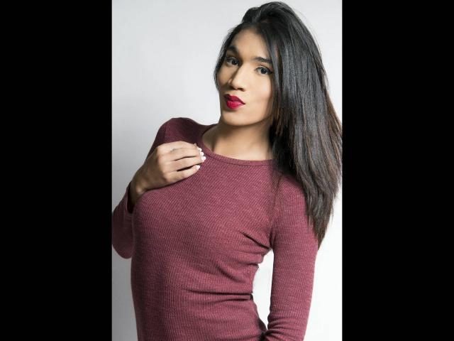 Littlebrunettehot with dark, straight hair, red lipstick, and a tight, maroon rib-knit top, hand on chest, looking at camera, medium breasts, light brown skin, standing.