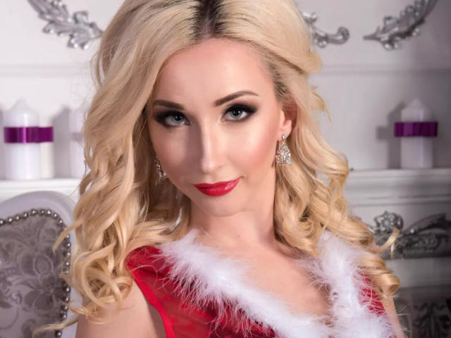 FrancescaHottyX met blonde krullen, rode lippenstift en een rode kanten jurk met witte bontrand, draagt diamanten oorbellen en glimlacht verleidelijk. Borsten zichtbaar onder het kant..