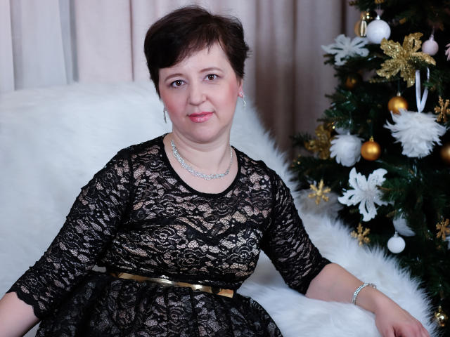 ChristaRose, a chubby woman with short brown hair, wearing a black lace dress, sitting on a white furry couch, next to a decorated Christmas tree. Her tits are small.