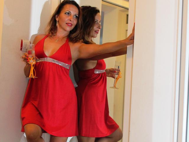 Aprel, met een gemiddelde borstmaat, in een rode jurk en een zilveren riem, houdt een glas champagne vast, leunt tegen een spiegel en glimlacht. Haar reflectie toont haar sensuele pose..