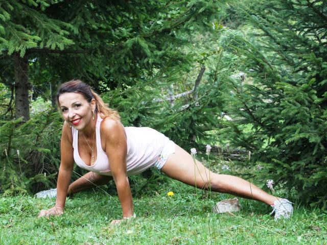 Aprel è a quattro zampe, sta facendo un plank in una foresta, indossa una canottiera rosa e pantaloncini colorati, con rossetto rosso e un sorriso, mostrando i suoi seni e i suoi glutei..