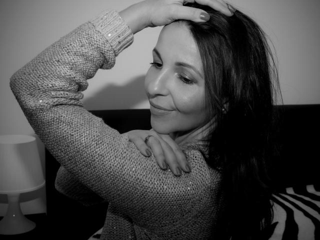 Aprel ha i capelli lunghi e scuri, indossa un maglione lavorato con una trama particolare, sorride con la mano tra i capelli, mostrando un anello al dito, e ha un bel sedere. Foto in bianco e nero..