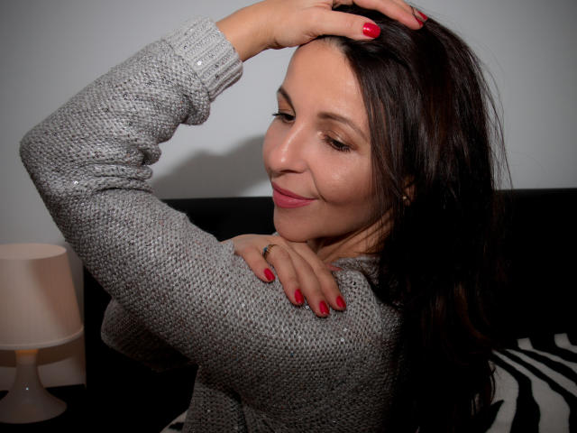 Aprel, con cabello oscuro, uñas rojas y un suéter de punto gris, sonriendo mientras se toca el cabello, mostrando pequeños senos y un rostro lindo. Iluminación suave, almohada con estampado de cebra..