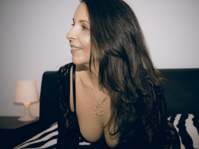 Aprel, con cabello largo y castaño, vestida con una blusa negra de encaje que muestra su escote, sonriendo, mirando hacia un lado, llevando un collar con forma de corazón, junto a una lámpara en una cama con estampado de cebra..
