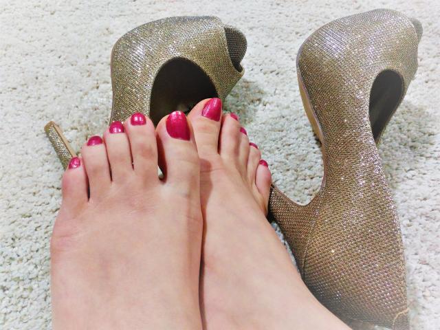 I piedi di GinnyGold, con l'unghie laccate di un rosa brillante, sono accanto a un paio di tacchi alti dorati scintillanti su un tappeto bianco. Le sue dita dei piedi sono leggermente incurvate, mettendo in mostra le sue unghie curate..