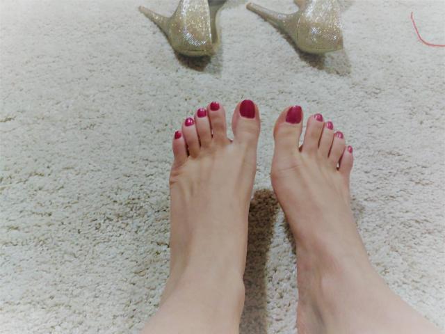 GinnyGolds Füße mit rot lackierten Zehen, stehend auf einem Teppich, mit glitzernden, beige farbenen Absätzen im Hintergrund..