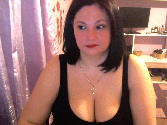 HellenBooty69 mit schwarzem Bob, rotem Lippenstift, schwarzem Tanktop, großen Brüsten, einer Halskette mit einem Kreuz, sitzend, leicht zur Seite blickend, in einem Raum mit violetten Vorhängen..