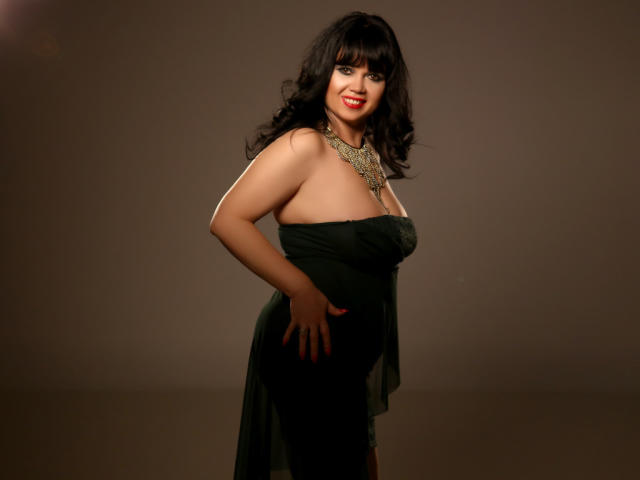 MatureVivian with dark curly hair, red lipstick, and a green, strapless dress, showcasing her large tits and curvy ass, wearing a gold necklace, smiling confidently.