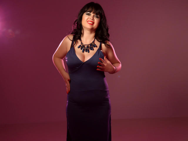 MatureVivian has dark wavy hair, red lipstick, and wears a navy blue dress with a deep cleavage, showing her big tits, and a blue necklace. She stands smiling.