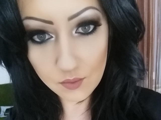 BeauxYeuxx a les cheveux noirs et ondulés, un maquillage des yeux fumé et des lèvres roses. Elle regarde l'appareil photo avec un léger sourire confiant, mettant en valeur son visage attrayant et séduisant..
