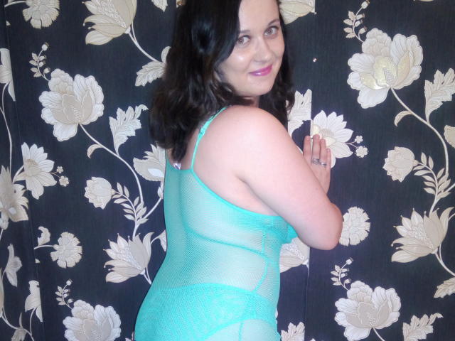 ChaudeBella69, avec sa peau pâle et ses cheveux noirs ondulés, porte une lingerie turquoise en dentelle transparente, elle sourit à la caméra, montrant son dos et ses fesses, ses mains pressées contre le mur..