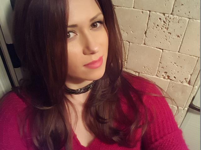 ToiSecret, avec ses cheveux bruns, son rouge à lèvres rose et son pull rouge, porte un collier noir, a un sourire discret et une peau claire, avec une légère inclinaison de la tête..