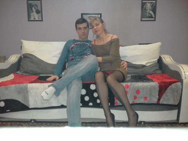 Bobandchris69, avec des seins, portant une robe marron à une épaule et des bas noirs, est assise sur un canapé. Bobandchris69, avec ses fesses, porte un jean bleu et une chemise turquoise..