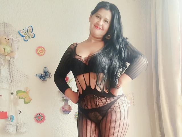 KinkyViolet está de pie, sonriendo, vestida con un conjunto de lencería negra transparente a rayas que deja ver sus grandes y redondos senos y revela sus bragas negras de encaje. Su largo cabello negro cae sobre un hombro..