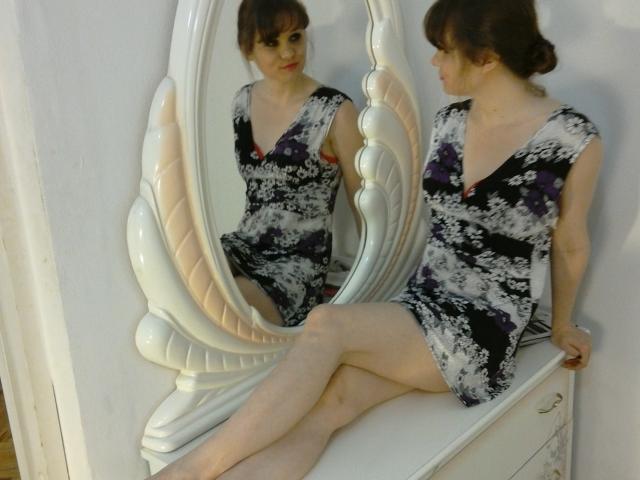 SassyCabotCaboche está sentada em uma cômoda branca, com as pernas cruzadas, vestindo um vestido preto e branco com detalhes roxos, refletindo em um espelho ornamentado, que mostra suas nádegas e seios..