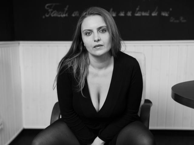 Oliviaaa with medium tits, sitting, wearing a black deep V-neck top, long hair, and black tights, looking at camera, in a black and white photo.