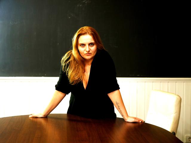 Oliviaaa with long red hair, black V-neck shirt, tattoo on right forearm, leaning on wooden table, arms spread, intense expression, white chair to her right.
