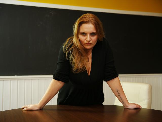 Oliviaaa, avec des cheveux roux, un t-shirt noir, appuyée sur une table en bois, les bras ouverts, un tatouage sur l'avant-bras droit, une expression sérieuse, une poitrine de taille moyenne, une chaise blanche, un mur jaune et noir..