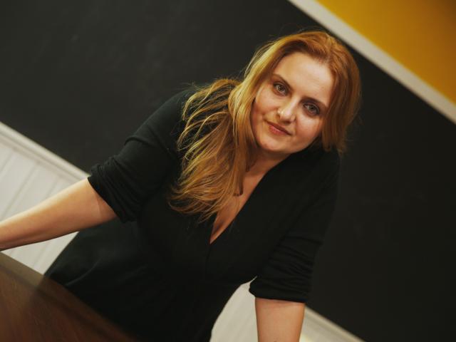 Oliviaaa, avec des cheveux roux, une robe noire et un décolleté, appuyée sur une table, souriant légèrement, dans une pièce faiblement éclairée. Ses fesses sont partiellement visibles..