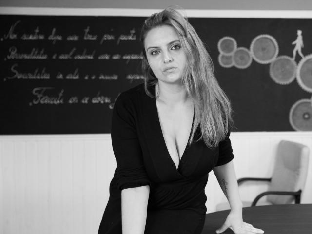 Oliviaaa, avec une poitrine de taille moyenne, porte un haut noir à col en V profond, elle est appuyée contre une table, avec une expression sérieuse, cheveux blonds, photo en noir et blanc, un tableau noir en arrière-plan, une chaise à droite..